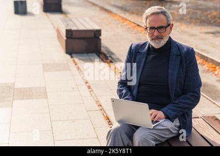 Remote Working-Konzept. Erwachsener kaukasischer grauhaariger bärtiger Geschäftsmann in formeller Kleidung profitiert von schönem sonnigen Herbstwetter und arbeitet draußen mit seinem Laptop. Blick auf die Kamera. Hochwertige Fotos Stockfoto