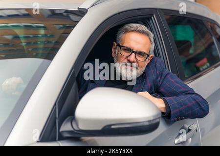 Hübscher und selbstbewusster bärtiger grauhaariger reifer Mann, der auf dem Fahrersitz in seinem Auto sitzt, sich aus dem Fenster lehnt und die Kamera anschaut. Hochwertige Fotos Stockfoto