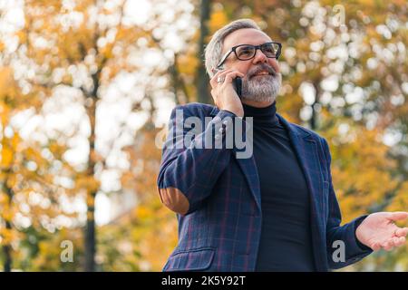 Kunde ruft beim 60-jährigen kaukasischen Firmenbesitzer an. Business-Freizeitkleidung. Telefongespräch. Moderne Technologie. Mittlere Nahaufnahme. Außenaufnahme. Unscharfe Bäume mit gelben Blättern im Hintergrund. Hochwertige Fotos Stockfoto