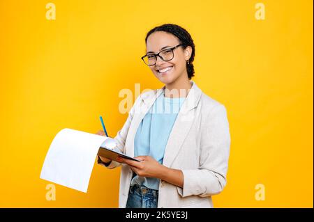 Schöne junge Brünette gemischt Rennen Frau, trägt stilvolle Freizeitkleidung, hr-Manager, Immobilienmakler, hält Ordner mit Papieren Dokument, schaut auf Kamera und lächeln, isoliert orange Hintergrund Stockfoto