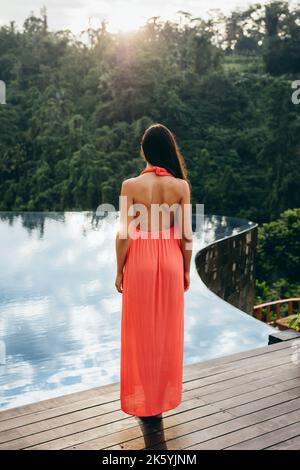Rückansicht eines weiblichen Models in der Sundress, das am Rand des Schwimmbades steht. Junge Frau am Pool eines Luxusresorts. Stockfoto