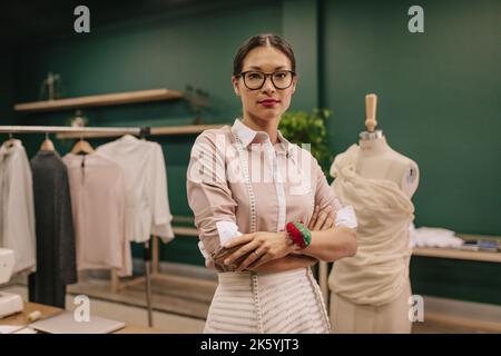 Erfolgreiche Designerin mit Maßband um den Hals, die in ihrer Werkstatt steht. Frau, die im Modestudio mit gekreuzten Armen steht. Stockfoto