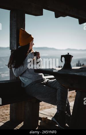 Frau mit einer Tasse Kaffee sitzt vor dem Blick auf die Berge und schaut weg Stockfoto
