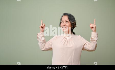 Schöne und glückliche 60s-jährige Asiatin lächelt, hebt ihre Finger oder zeigt nach links und rechts Finger nach oben, isoliert auf grünem Hintergrund stehend. Stockfoto