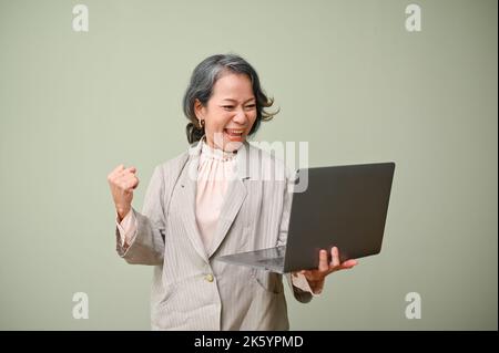 Aufgeregt und fröhlich Senior asiatische Geschäftsfrau hält und Blick auf ihren Laptop-Bildschirm, hebt ihre Hand, schreiend vor Glück. Ja, wir feiern vi Stockfoto