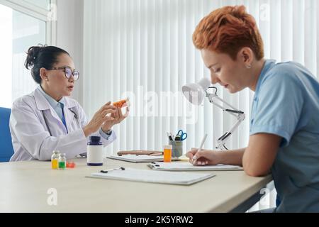 Junge Krankenschwester, die am Tisch ein Rezept schreibt, wobei der Arzt vor ihr sitzt und Pillen untersucht Stockfoto