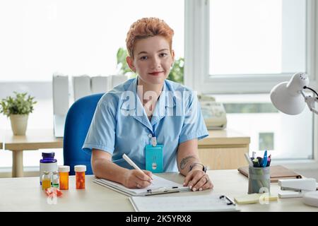 Porträt einer jungen Krankenschwester, die auf die Kamera schaut, während sie am Tisch ein Rezept auf die Patientenkarte schreibt Stockfoto