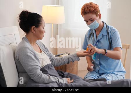 Junge Krankenschwester in Uniform, die einer kranken älteren Frau Pille gab, während sie im Bett saß Stockfoto