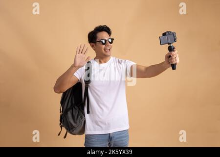 Junger asiatischer Tourist Backpacker Mann lächelt und nimmt ein Selfie und Blog isoliert auf beigem Hintergrund Stockfoto