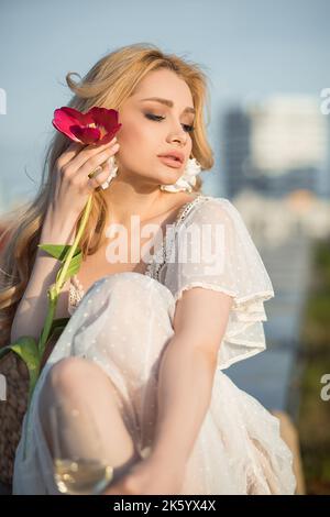 Schöne junge Frau in hübschen Kleid und Ohrringe sitzen auf der Dachterrasse mit rosa Tulpe Nahaufnahme. Foto von pariser Dame auf Himmel Hintergrund. Paris Stockfoto