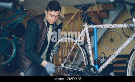 Erfahrener Mechaniker in Handschuhen montiert das Hinterrad des Fahrrads während der Wartung des Fahrzeugs am Arbeitsplatz. Professionelle Instrumente, Ersatzteile und Geräte sind sichtbar. Stockfoto