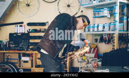 Ein erfahrener Wartungstechnischer arbeitet an seinem kleinen Arbeitsplatz mit einer elektrischen Kreissäge. Der junge Mann trägt Schutzbrille und Handschuhe und hört mit Ohrhörern Musik. Stockfoto