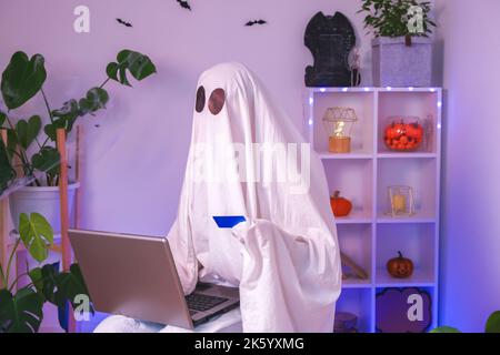 ghost of Halloween nutzt Laptop im Internet surfen, Online-Shops durchsuchen, Märkte. Ein Geist macht eine Bestellung vom Computer für Halloween. Ghost zahlt für den Online-Kauf per Kreditkarte. Neonlicht. Stockfoto