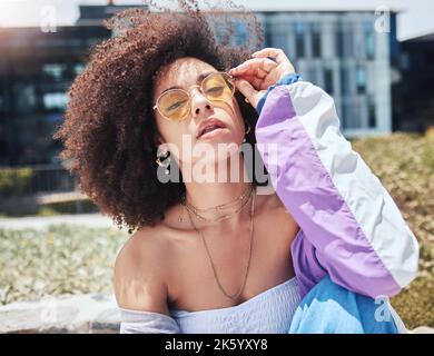 Junge stilvolle Mixed-Race-Frau mit lockigen natürlichen Afro-Haaren trägt trendige Sonnenbrille und bunte Vintage-Jacke. Ein Weibchen sieht nur cool aus und Stockfoto