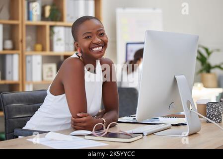 Fröhliche junge afroamerikanische Geschäftsfrau, die lächelt und in einem Büro am Computer arbeitet. Kahle Unternehmerin, die selbstbewusst und glücklich ist Stockfoto