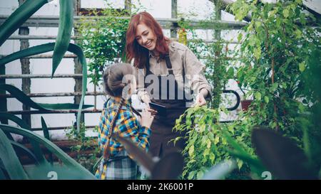 Junge Mutter und Tochter Moderne Bauern reden, lachen und haben Spaß, während sie Pflanzen im Gewächshaus kontrollieren. Familienunternehmen, Agrarkultur und Menschen Konzept. Stockfoto