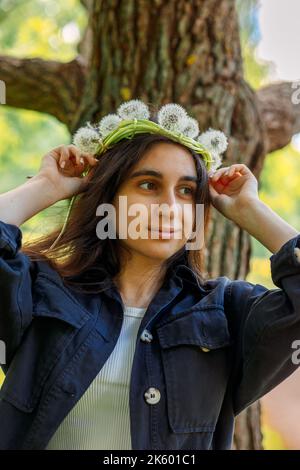 Glückliches Mädchen mit Kranz aus weißem Elendelion auf grünem Rasen am sonnigen Sommertag. Hochwertige Fotos Stockfoto