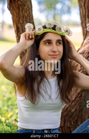 Glückliches Mädchen mit Kranz aus weißem Elendelion auf grünem Rasen am sonnigen Sommertag. Hochwertige Fotos Stockfoto