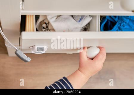 Das Baby öffnet das Kinderschloss in der geschlossenen Schublade des Schranks. Kleinkind Baby mit Hand öffnet Türschloss auf der Brust der Schubladen. Kind im Alter von einem Jahr und drei Stockfoto