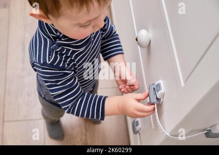 Das Baby öffnet das Kinderschloss in der geschlossenen Schublade des Schranks. Kleinkind Baby mit Hand öffnet Türschloss auf der Brust der Schubladen. Kind im Alter von einem Jahr und drei Stockfoto