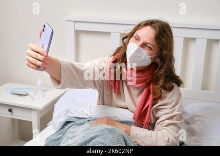 Kranke Bloggerin macht Selfie-Foto auf dem Handy Apple iPhone. Erwachsene kranke Frau in einem roten Schal gewickelt sitzt auf einem Heimbett in einem weißen Schlafzimmer, weibliches Alter Stockfoto