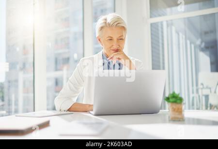 Eine fokussierte, reife kaukasische Geschäftsfrau, die mit der Hand auf dem Kinn denkt, während sie in einem Büro an einem Laptop arbeitet. Nur eine Frau plant und erwägt Stockfoto