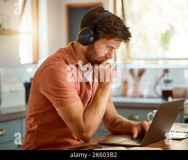Junger, ernsthafter kaukasischer Geschäftsmann, der Kopfhörer trägt und Musik hört, während er alleine zu Hause einen Laptop benutzt. Ein auf Männer ausgerichtetes Unternehmen Stockfoto