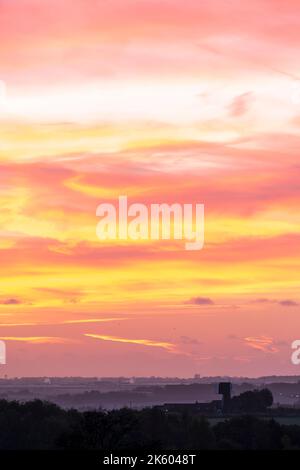 Der Morgenhimmel über der Landschaft von Kent blickt auf die Isle of Thanet. Verschiedene Schichten von Feldern, Bäumen und Hecken auf einer sanft rollenden, aber überwiegend flachen Landschaft. Himmel mit einigen verstreuten Wolkenschichten, lebhafter Gelb- und Rotfärbung. Stockfoto