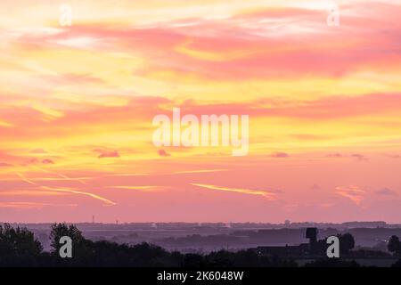 Der Morgenhimmel über der Landschaft von Kent blickt auf die Isle of Thanet. Verschiedene Schichten von Feldern, Bäumen und Hecken auf einer sanft rollenden, aber überwiegend flachen Landschaft. Himmel mit einigen verstreuten Wolkenschichten, lebhafter Gelb- und Rotfärbung. Stockfoto