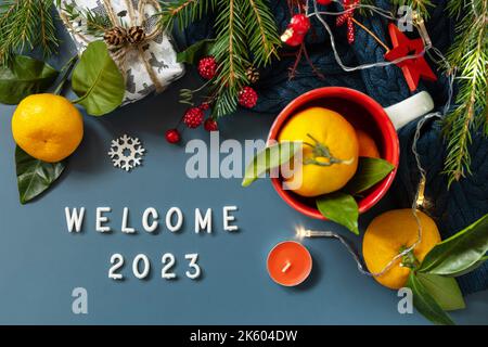 Begrüßungstext 2023. Winterkomposition mit Tannenzweigen, Gegenwart, Zapfen und Mandarinen auf blauem Hintergrund. Gemütliche Winterstimmung. Blick von oben. Stockfoto