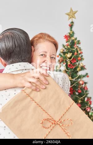 Ältere lateinische Paare feiern die Feiertage gemeinsam zu Hause, sie umarmen sich glücklich, während sie Weihnachtsgeschenke austauschen, sie schaut auf die Kamera, während sie Stockfoto