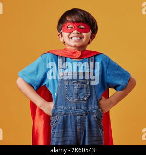 Entzückender kleiner asiatischer Junge mit Maske und Umhang, der sich vor einem orangen Studiohintergrund als Superheld ausgibt. Netter glücklicher Junge, der vorgibt Stockfoto