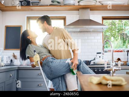 Fröhliches junges interrassisches Paar, das tanzt, während es gemeinsam ihr Zuhause putzt. Junger kaukasischer Mann und hispanische Frau, die Spaß haben, während sie Hausarbeiten machen Stockfoto