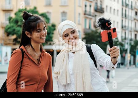 Multiethnische Freundinnen, die lächeln, während sie mit einem Mobiltelefon im Freien auf der Straße streamen oder aufzeichnen. Stockfoto