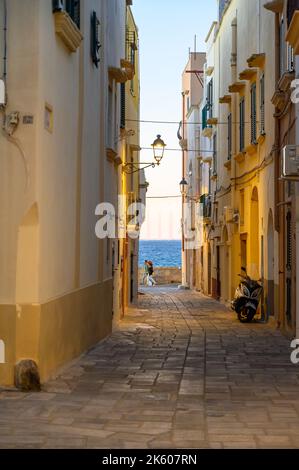 Ein Paar, das am Ende einer typischen engen und gepflasterten Straße in der Altstadt von Gallipoli, Apulien (Apulien), Italien, vorbeigeht. Stockfoto