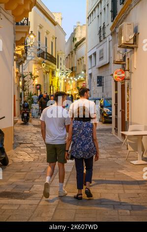 Touristen, die am frühen Abend in der Altstadt von Gallipoli, Apulien (Apulien), Italien, entlang einer typischen engen und gepflasterten Straße spazieren. Stockfoto