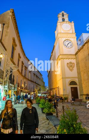 Touristen und Einheimische, die am Glockenturm der Kathedrale von Gallipoli auf einer gepflasterten Straße in der Altstadt von Gallipoli am Abend vorbeilaufen, Apulien (Apulien), Italien. Stockfoto