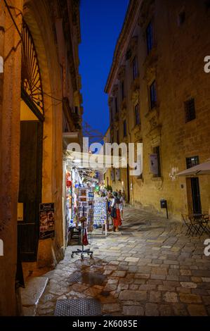 Touristen und Einheimische, die abends in der Altstadt von Gallipoli, Apulien (Apulien), Italien, eine typische enge und gepflasterte Straße entlang spazieren. Stockfoto