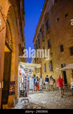 Touristen und Einheimische, die abends in der Altstadt von Gallipoli, Apulien (Apulien), Italien, eine typische enge und gepflasterte Straße entlang spazieren. Stockfoto