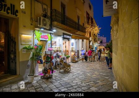 Touristen und Einheimische, die abends in der Altstadt von Gallipoli, Apulien (Apulien), Italien, eine typische enge und gepflasterte Straße entlang spazieren. Stockfoto