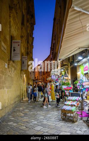 Touristen und Einheimische, die abends in der Altstadt von Gallipoli, Apulien (Apulien), Italien, eine typische enge und gepflasterte Straße entlang spazieren. Stockfoto