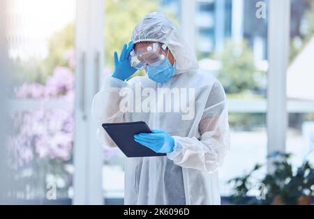 Gestresste und besorgte Frau, die ein digitales Tablet benutzt, während sie einen Schutzanzug mit Gefahrgut trägt, um sich im Krankenhaus oder Labor vor Krankheiten und Krankheiten zu schützen. Eins Stockfoto