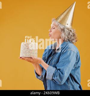 Eine glückliche reife kaukasische Frau bläst Kerzen auf einen Kuchen, den sie hält, während sie einen Geburtstagshut vor einem gelben Hintergrund im Studio trägt Stockfoto