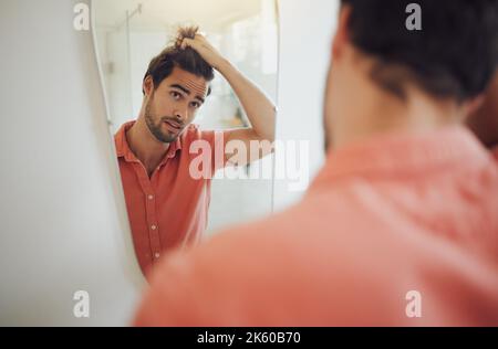 Ein hübscher junger Kaukasier, der seine Haare berührte und in den Badezimmerspiegel schaute. Mann zieht an den Haaren und denkt an einen Haarschnitt. Betroffen Stockfoto