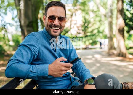 Bild eines glücklichen erwachsenen Mannes mit Sonnenbrille, der im Park auf der Bank sitzt und sein Telefon benutzt. Stockfoto