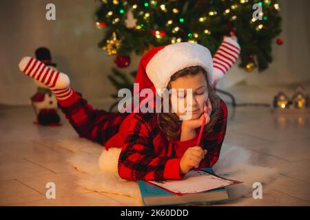 Ein kleines Mädchen in einem Weihnachtsmann-Hut schreibt einen Brief an den Weihnachtsmann zu Hause, der auf dem Bokeh-Boden im Hintergrund liegt. Winterkleidung für Weihnachten. Frohe Stockfoto