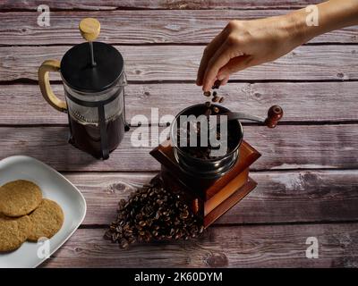 Lassen Sie Kaffeebohnen von Hand in eine Kaffeemühle neben einer Kaffeekocher und einem Teller mit Keksen fallen Stockfoto