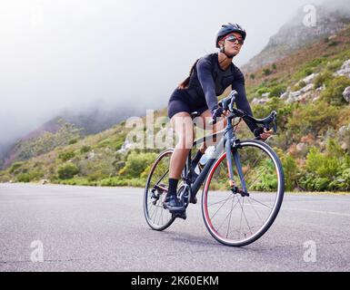 Eine athletische junge Frau, die draußen unterwegs ist. Sportlich-fitte Frau mit Helm und Brille beim Fahrradfahren auf einer Straße entlang des Berges zum Trainieren Stockfoto