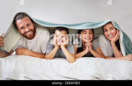 Porträt einer glücklichen vierköpfigen kaukasischen Familie, die in einer Reihe auf einem Bett mit einer Fleecedecke über dem Kopf liegt. Sorglose Eltern verbringen Freizeit mit Stockfoto