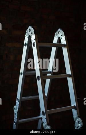 Konstruktion. Klappbare Leiter aus Metall in einem dunklen Raum auf einer Baustelle. Nahaufnahme der neuen Bauleiter auf Backsteinwand Hintergrund. Stockfoto